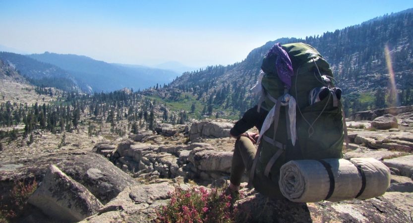 A person faces away from the camera while sitting on a rock, overlooking a mountainous landscape. 